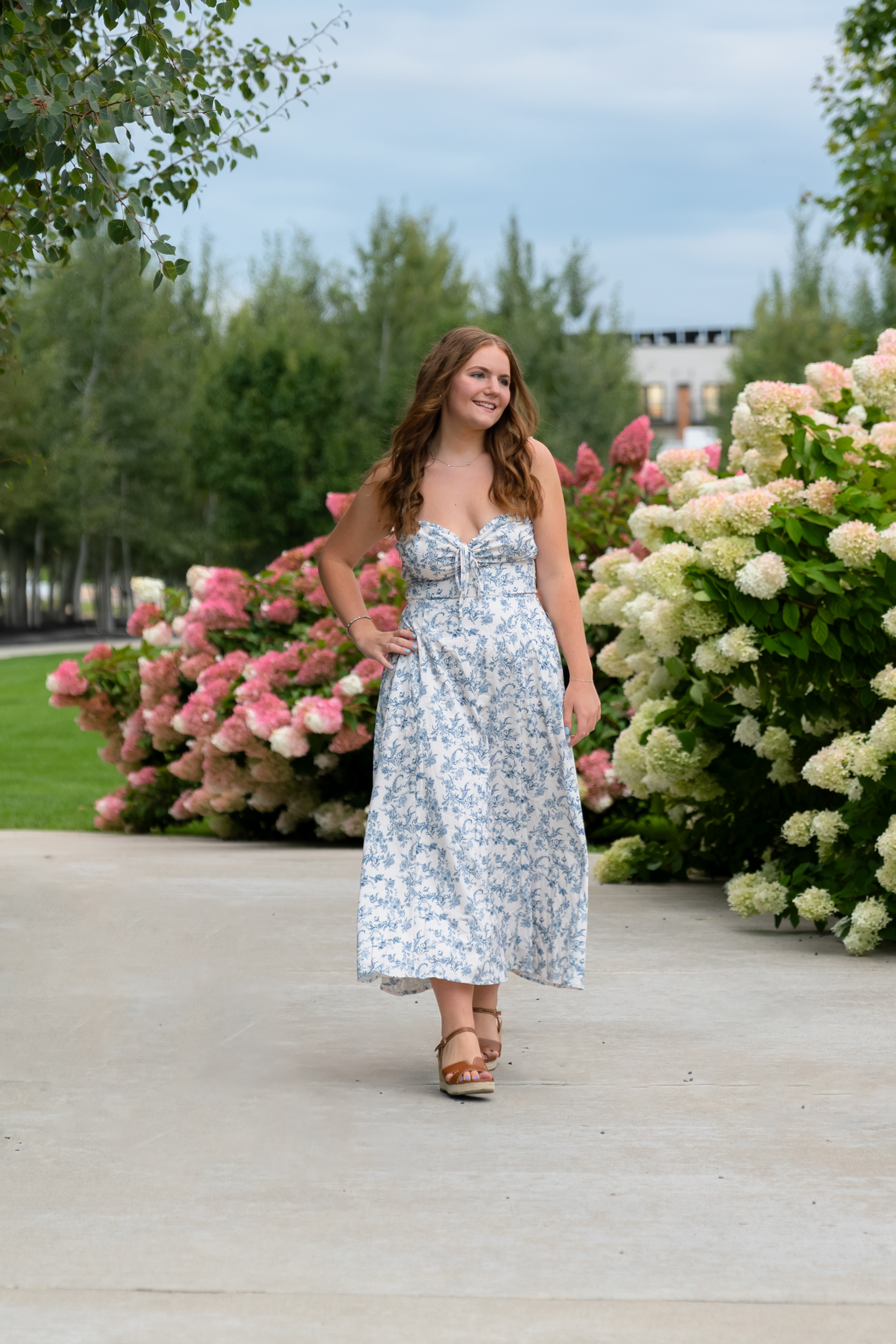 Young senior girl in flower print dress walking on a paved walking path between blooming flowers