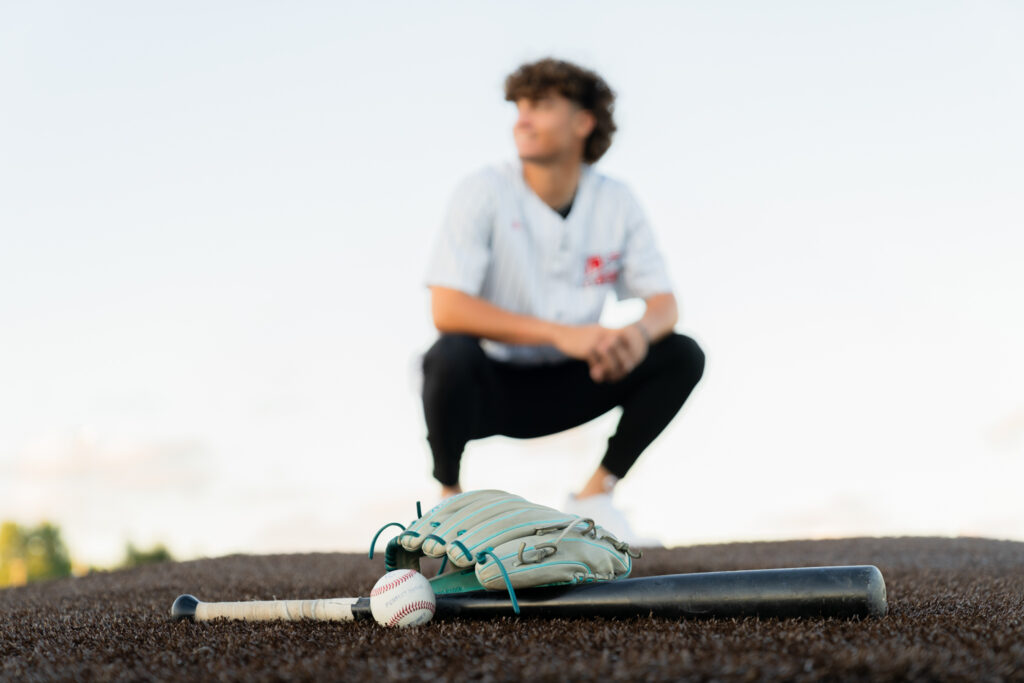 Boy squatting in his baseball uniform with his baseball gear in the foreground