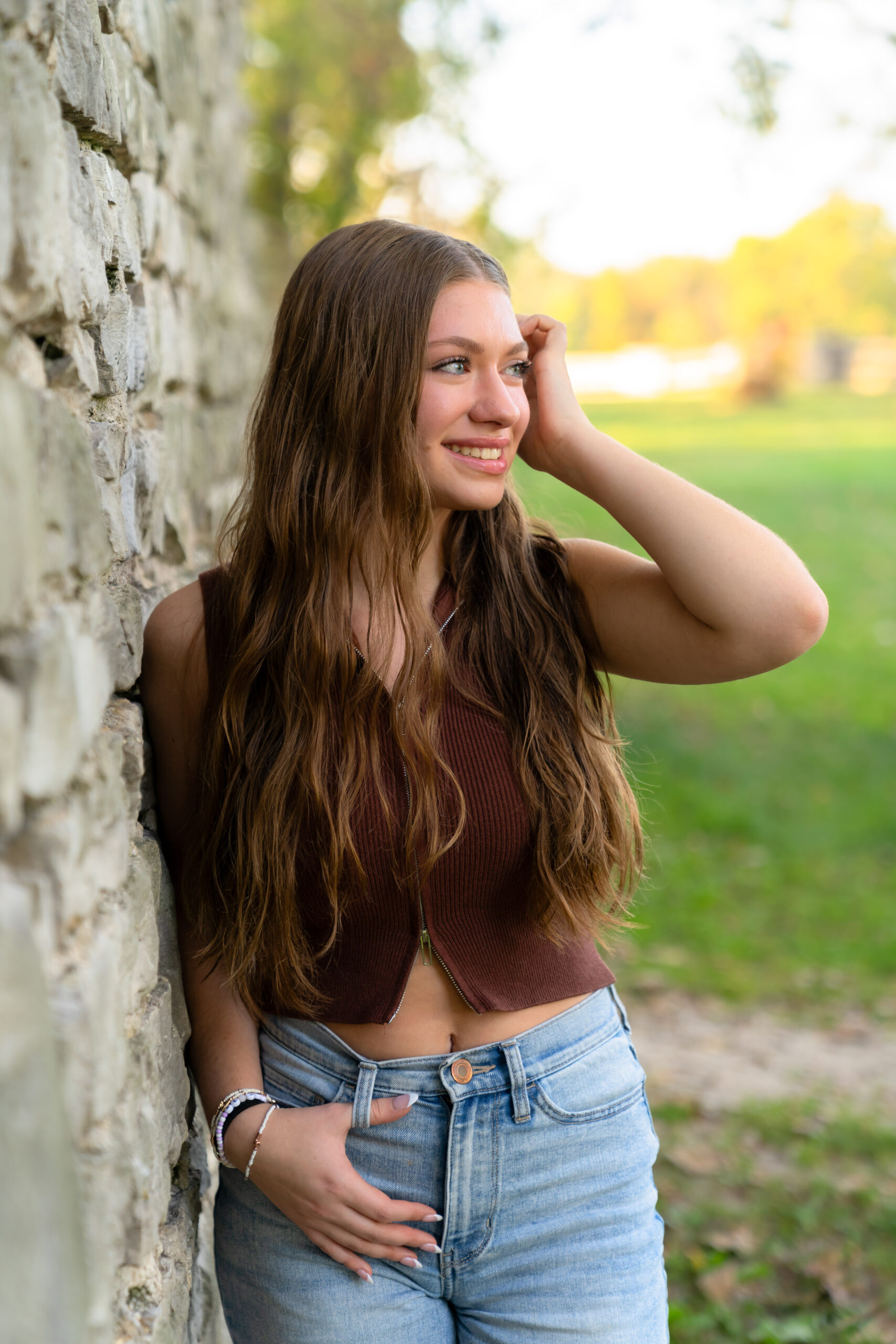 Senior girl in Lannon Stone quarry leaning against a brick wall with her hand looped through her jeans and tucking her brown hair behind her ear