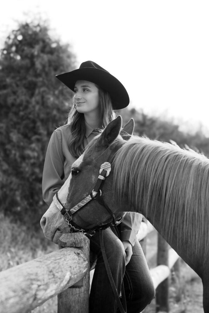 Cowgirl sitting on wooden fence with her horses head on her leg