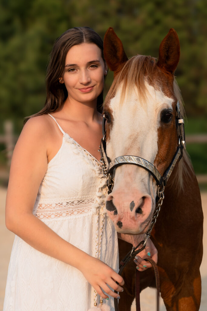 High school girl in white dress gently smiling at the camera with her horse