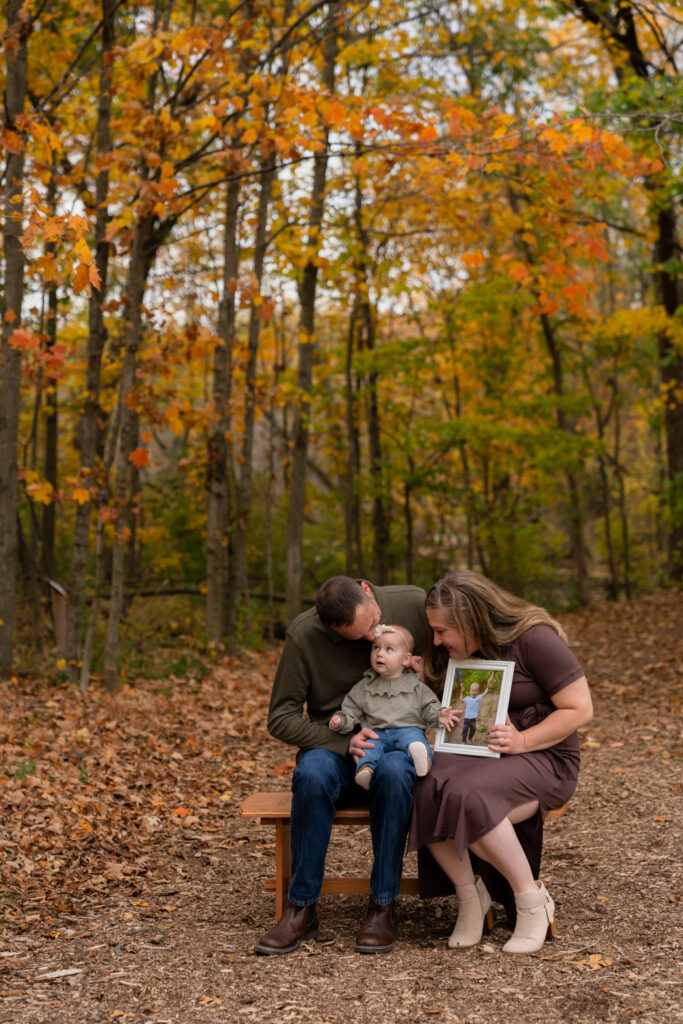 Family sitting on a bench in the woods during fall and smiling at one another while holding a framed photo of their son