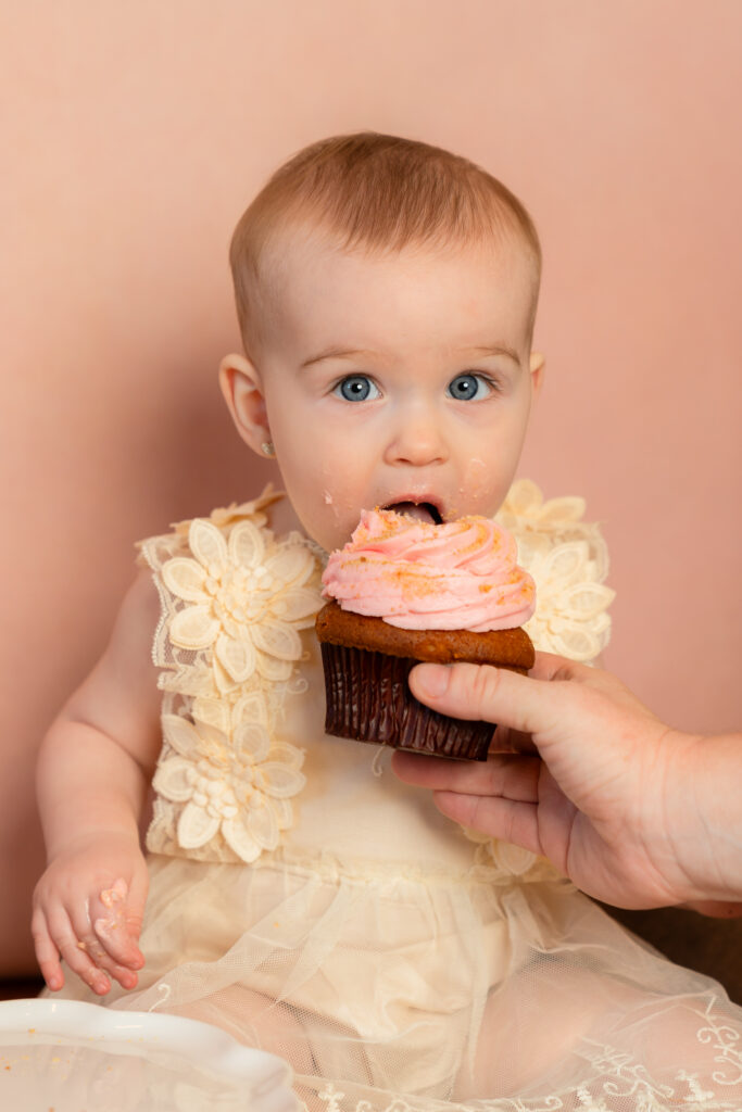 Little girl eating a cupcake while her mom holds it for her during cake smash in studio