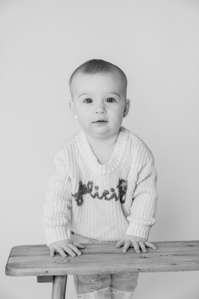 Little girl standing behind a wooden bench and looking at the camera during in-studio milestone session