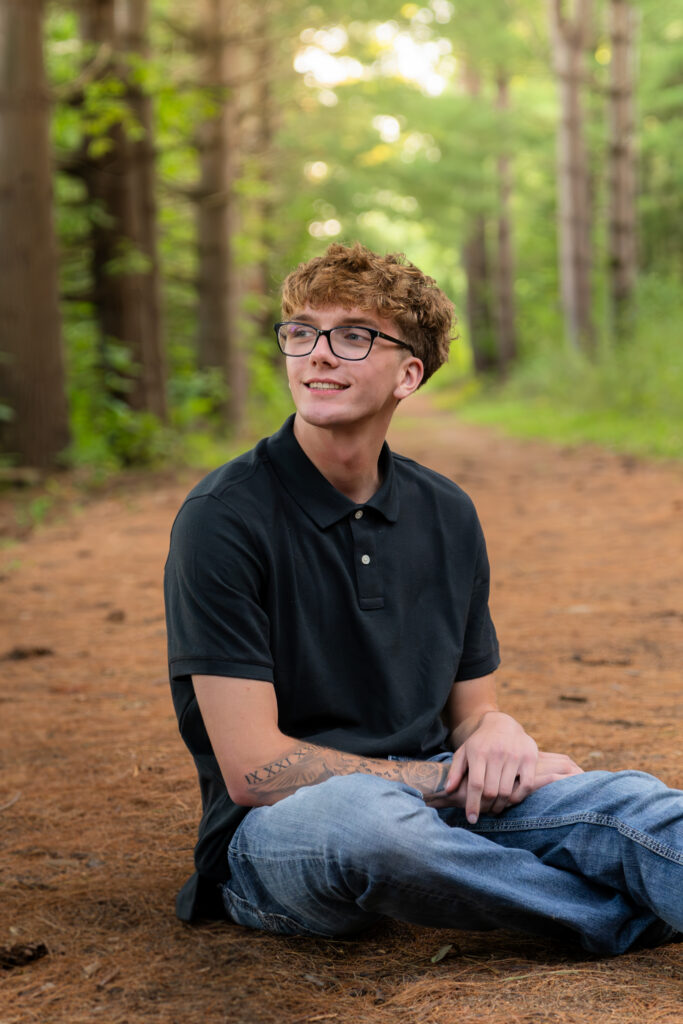High school boy with glasses sitting in tall pines at Leow Lake in Erin, wisconsin