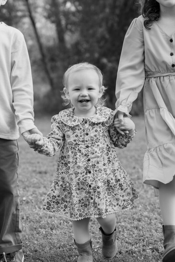Little girl holding her big brother and big sisters hand and smiling at the camera during family photo session