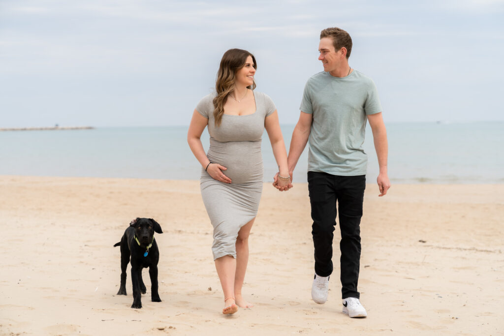 Couple holding hands and walking along the beach with their dog during maternity session in port Washington Wisconsin