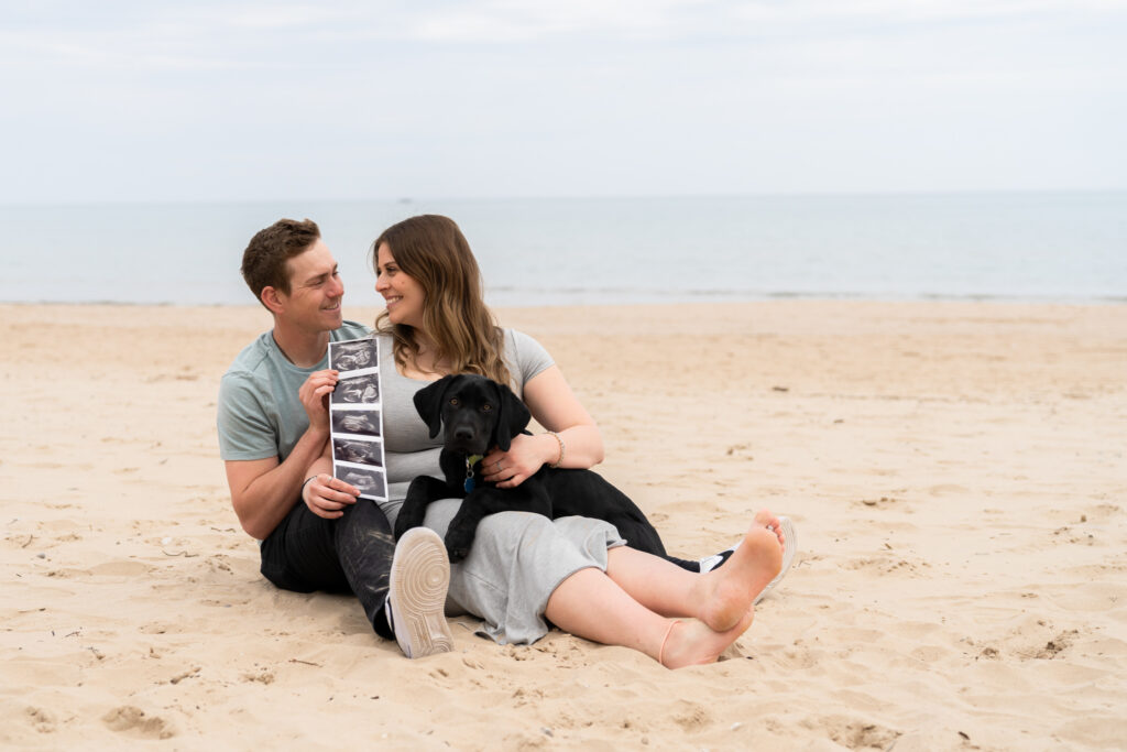 Man and woman holding sonogram ultrasound while sitting on the beach in Port Washington with their dog