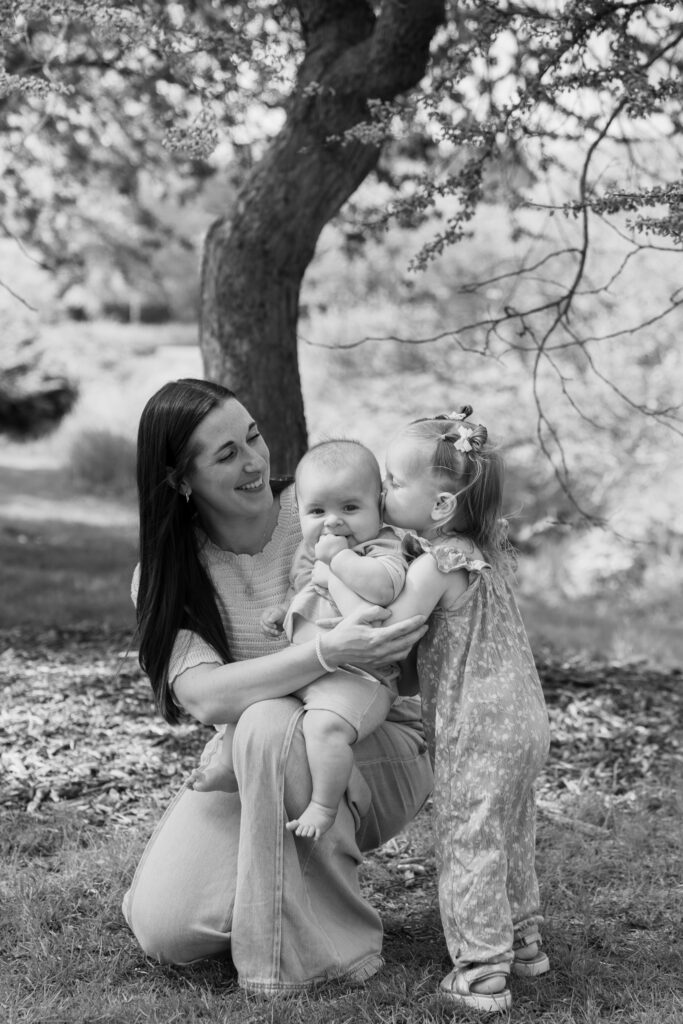 Big sister kissing her baby brother on the cheek while mommy holds him during family spring photography session