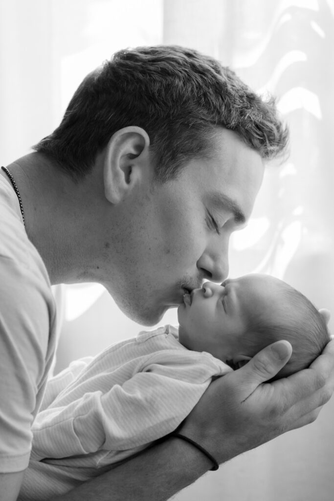 Dad kissing his newborn baby daughter in front of window during in-home newborn photography session