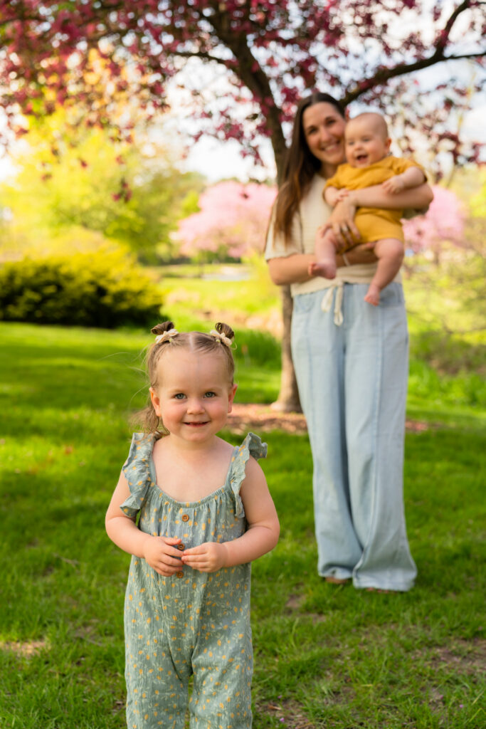 Little girl smiling at the camera while her mom holds her baby brother in the background in Waukesha, Wisconsin