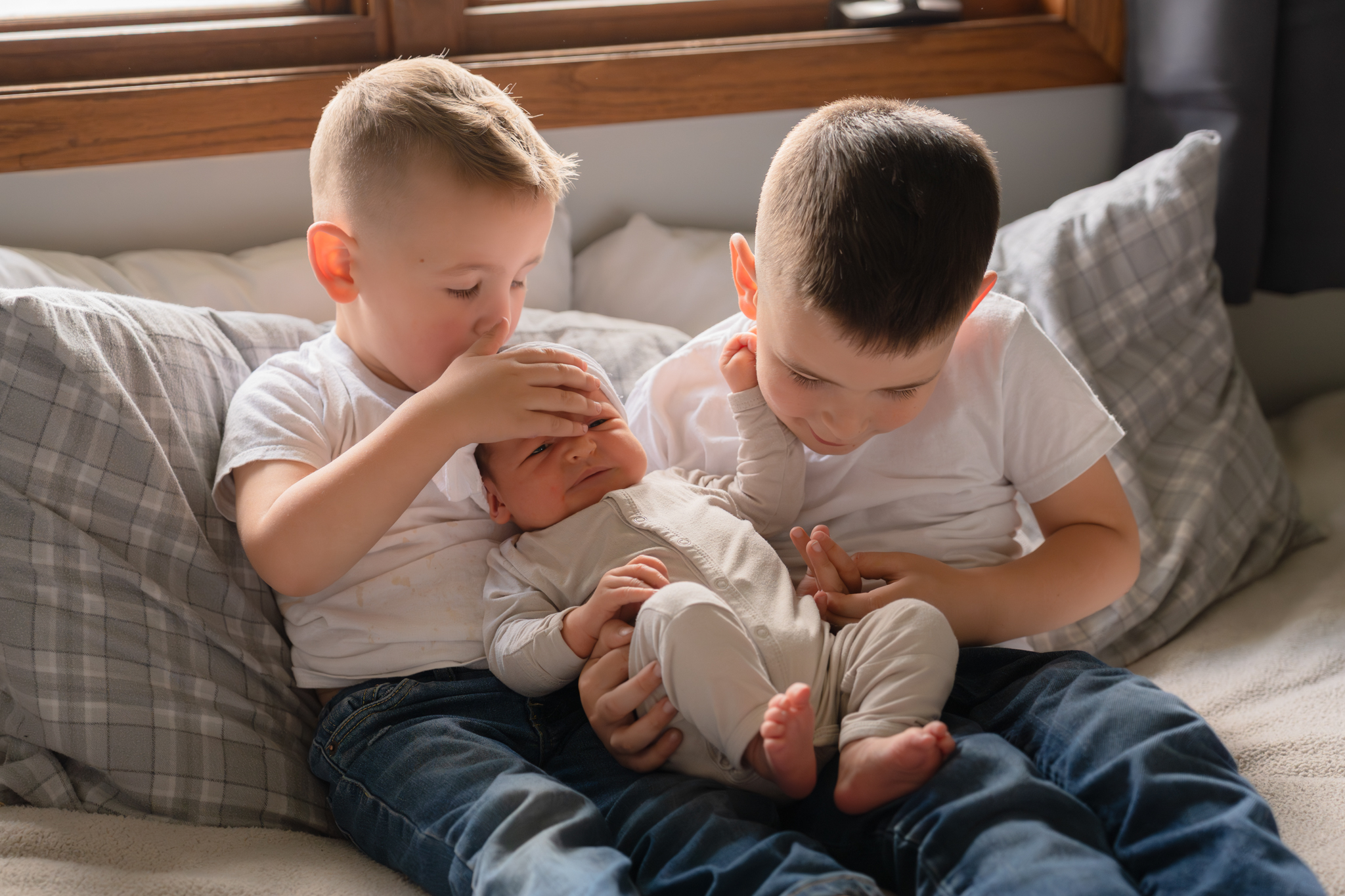 Big brothers holding their newborn baby sister while sitting on the bed and one of the boys kisses her on the top of her head