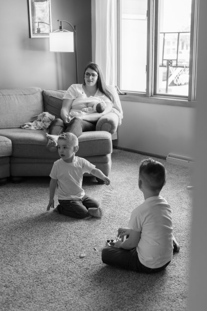 Mother breastfeeding her newborn baby on the couch while her two sons play on the floor in the living room