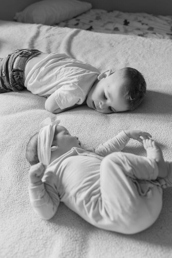 Little brother and his newborn baby sister laying on the bed and looking at each other while photographer takes a photo