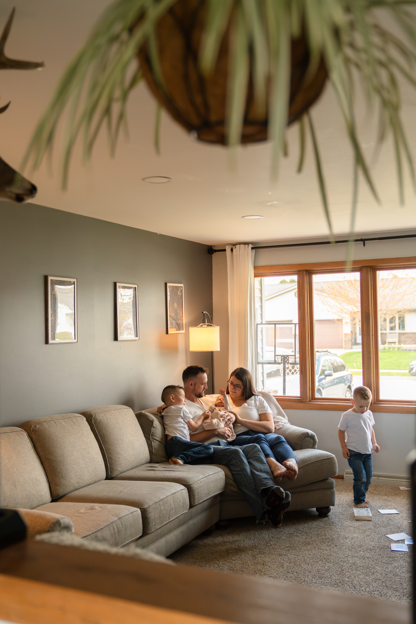 Family sitting on the couch in their living room and holding newborn baby while photographer captures the candid moment from afar