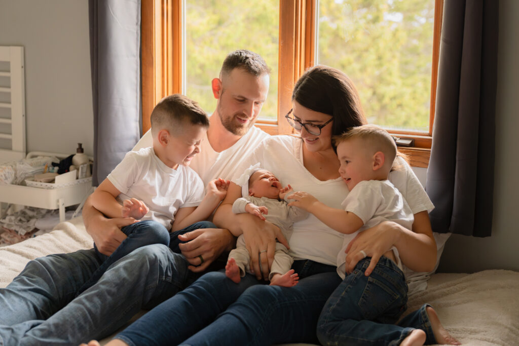 Mom and dad sitting on the bed with their two boys and newborn baby girl during cozy in-home newborn session with MelMarie Photography