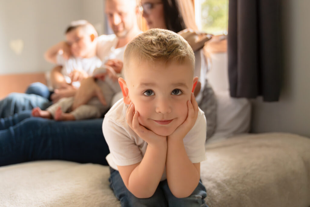 Young boy smiling at the camera with family sitting in the background during newborn photography session