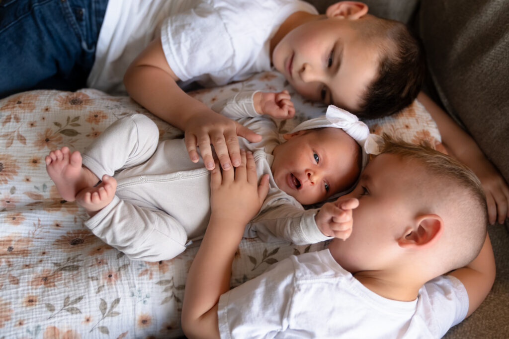 Two brothers laying next to their baby sister on the couch and smiling at her during newborn photo session
